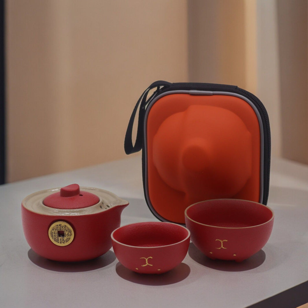 A compact red travel case opened to reveal a vibrant red ceramic teapot and two matching cups with gold lucky cat designs.
