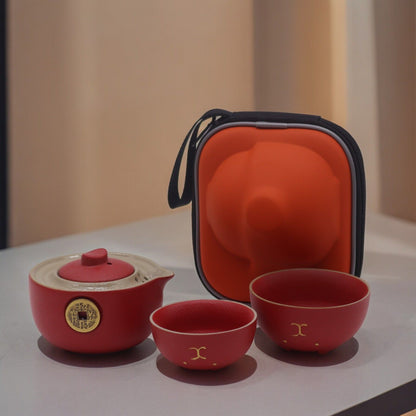 A compact red travel case opened to reveal a vibrant red ceramic teapot and two matching cups with gold lucky cat designs.
