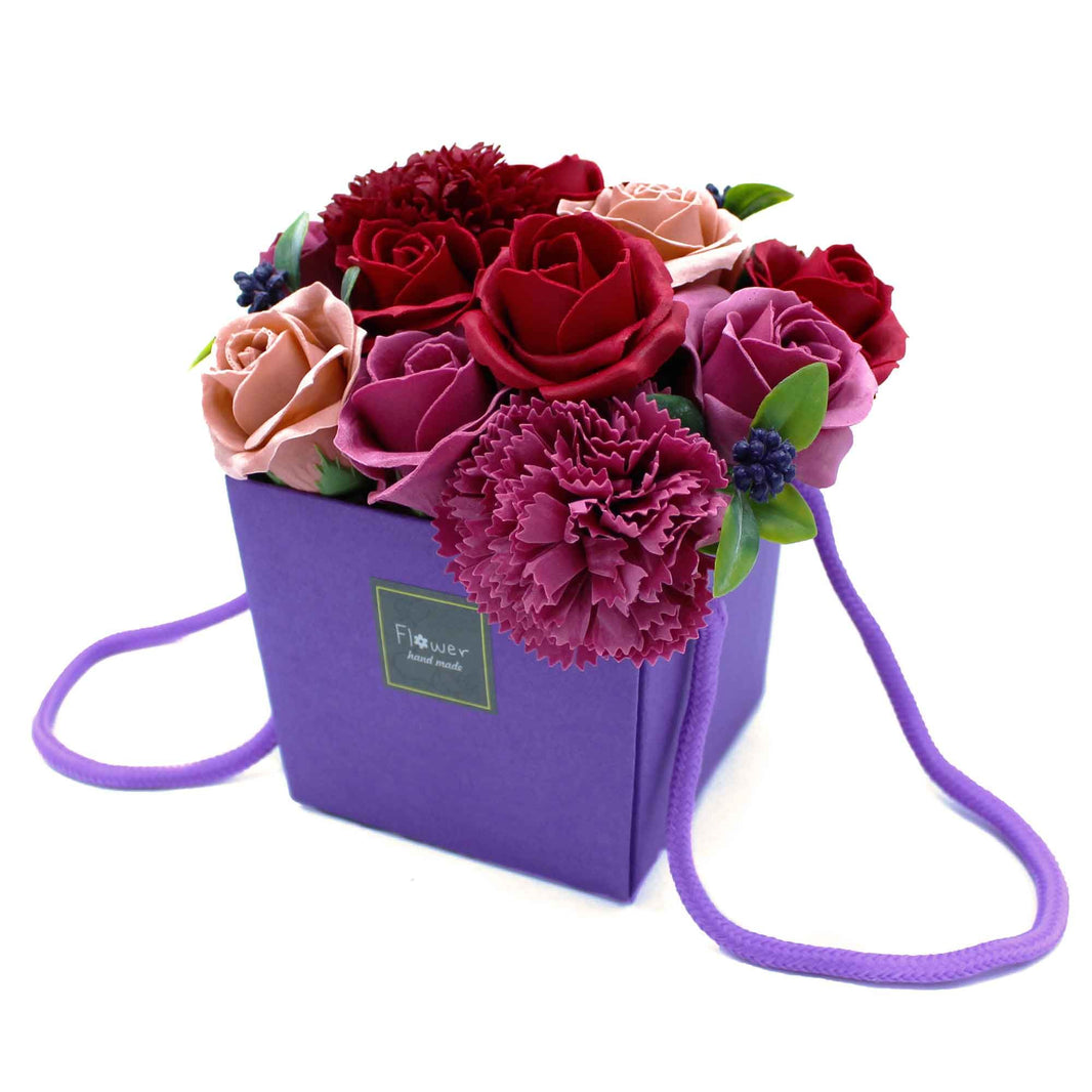 A vibrant purple soap flower bouquet with roses and greenery arranged in a decorative purple gift box with a carrying handle.