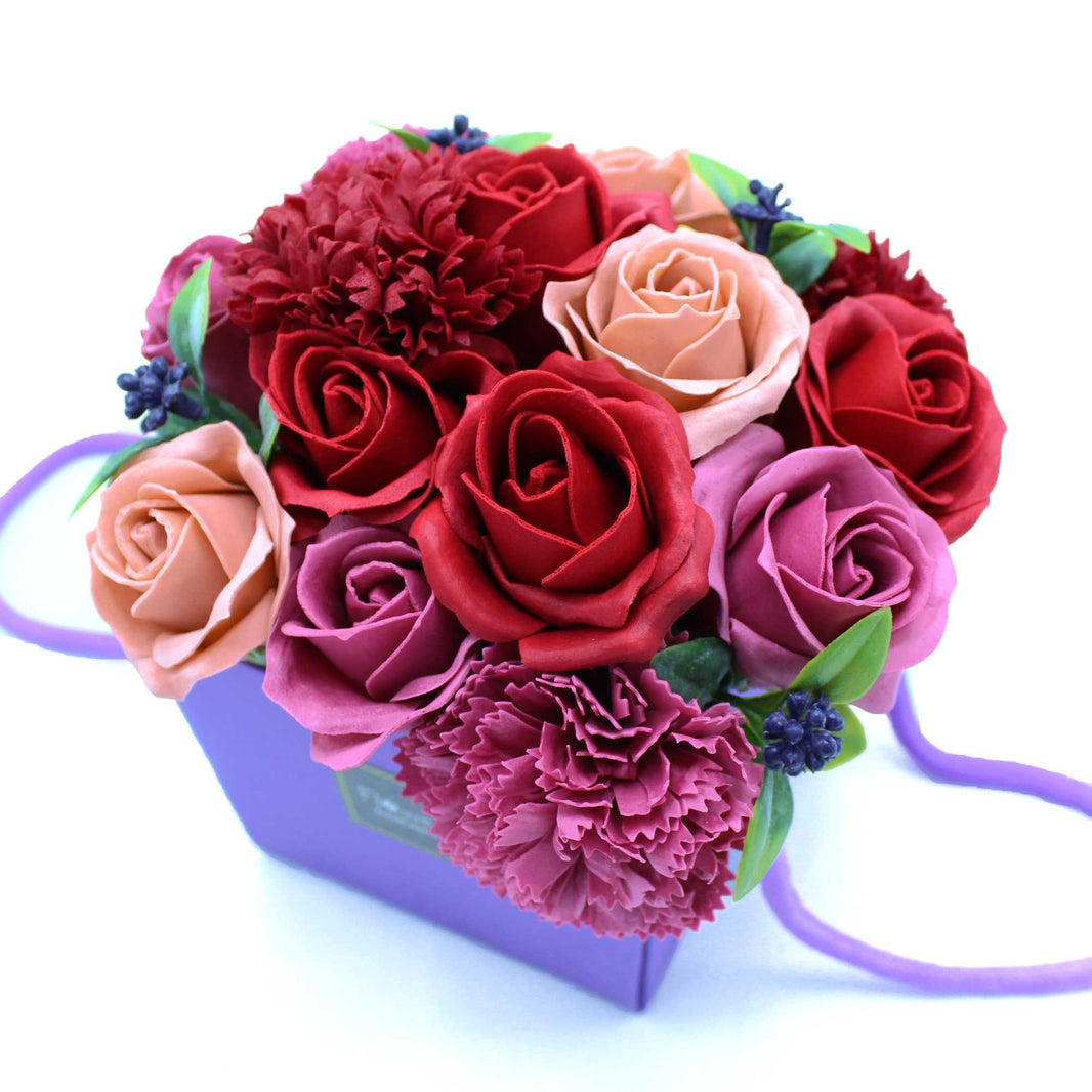 A vibrant purple soap flower bouquet with roses and greenery arranged in a decorative purple gift box with a carrying handle.