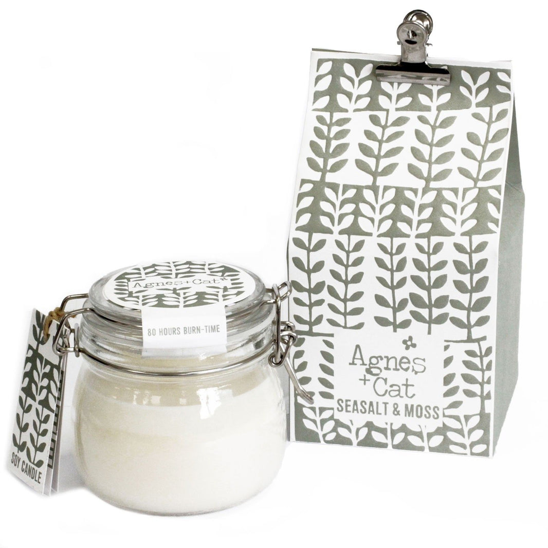 A classic clear glass Kilner jar filled with a white scented candle and a grey-toned decorative tag showing a mossy forest pattern.