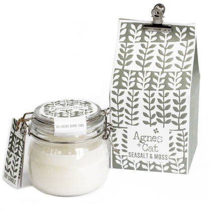 A classic clear glass Kilner jar filled with a white scented candle and a grey-toned decorative tag showing a mossy forest pattern.