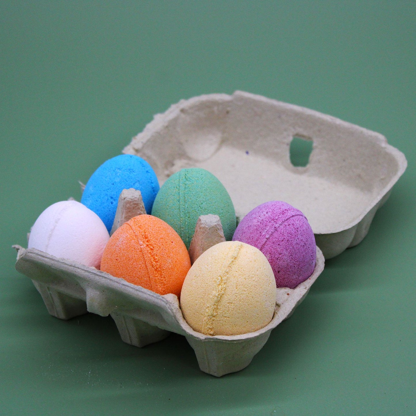 A cardboard egg carton filled with six different colored egg-shaped bath bombs, perfect for a Spring or Easter gift set.