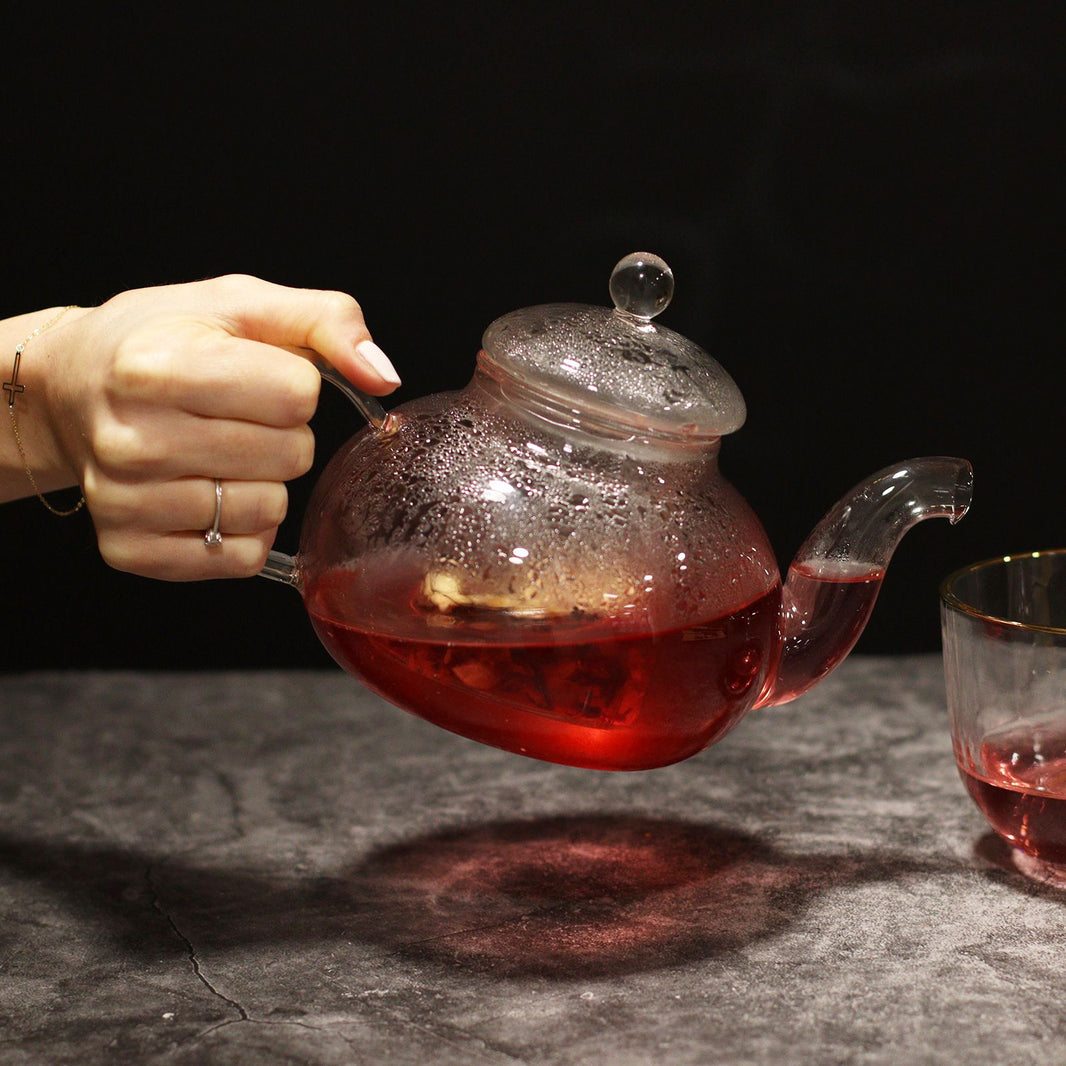 A clear, spherical borosilicate glass teapot with a fine mesh infuser, shown with dark red tea inside.