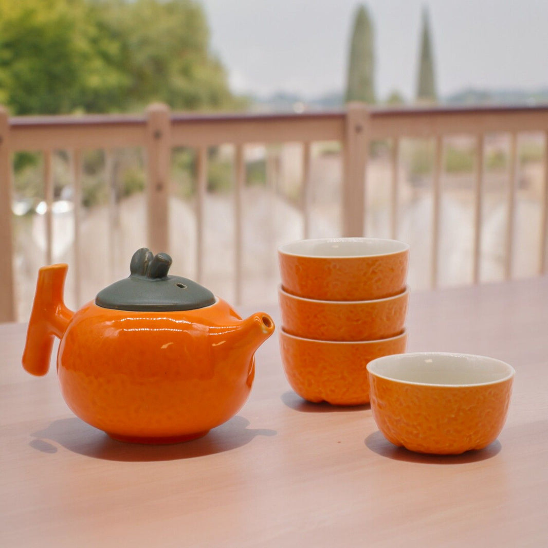 A contemporary orange ceramic teapot with four matching handleless cups, presented on a smooth wooden surface.