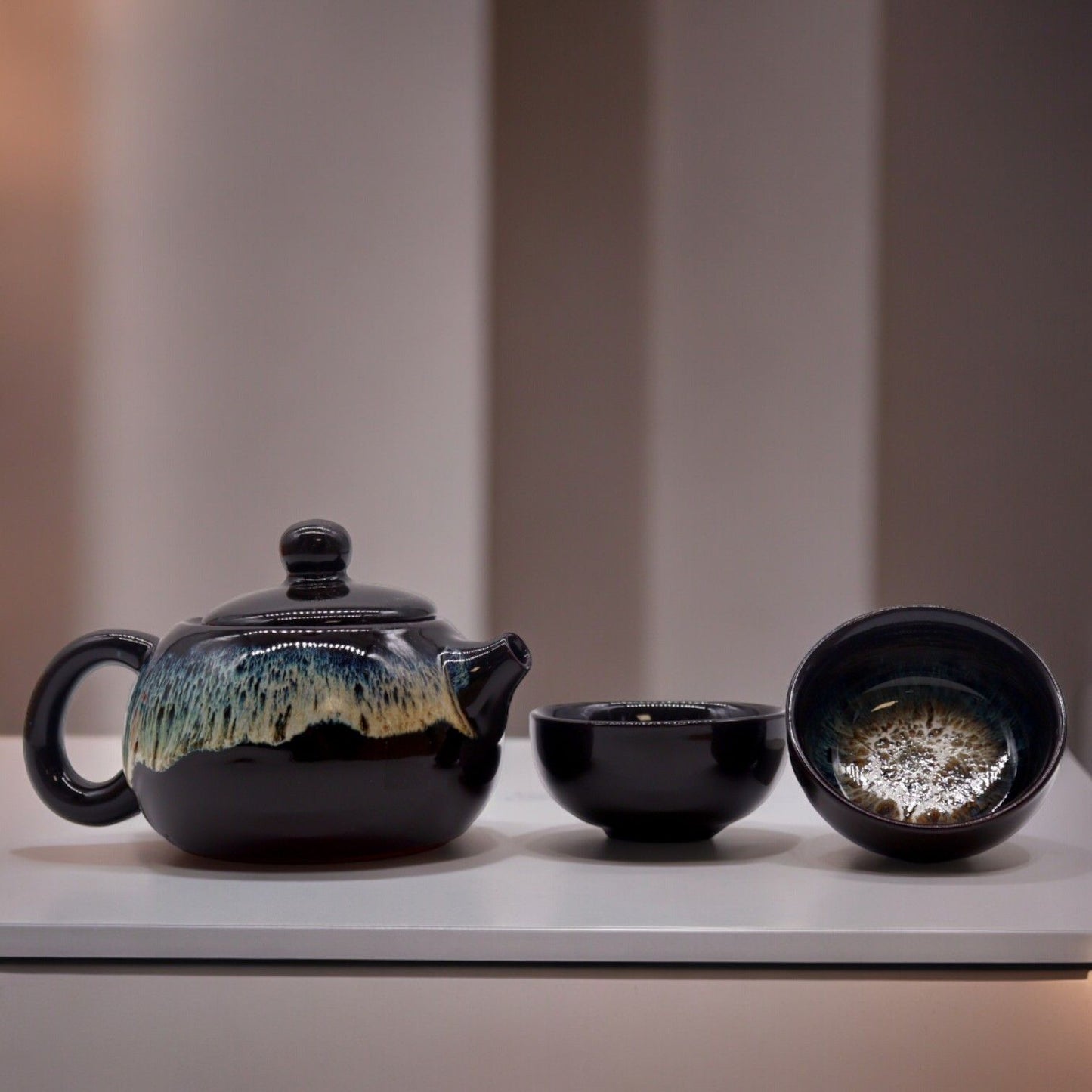A contemporary ceramic teapot set with a dark metallic black glaze, featuring a rounded pot and two matching handleless cups.