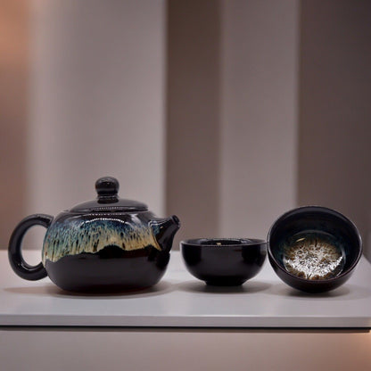 A contemporary ceramic teapot set with a dark metallic black glaze, featuring a rounded pot and two matching handleless cups.