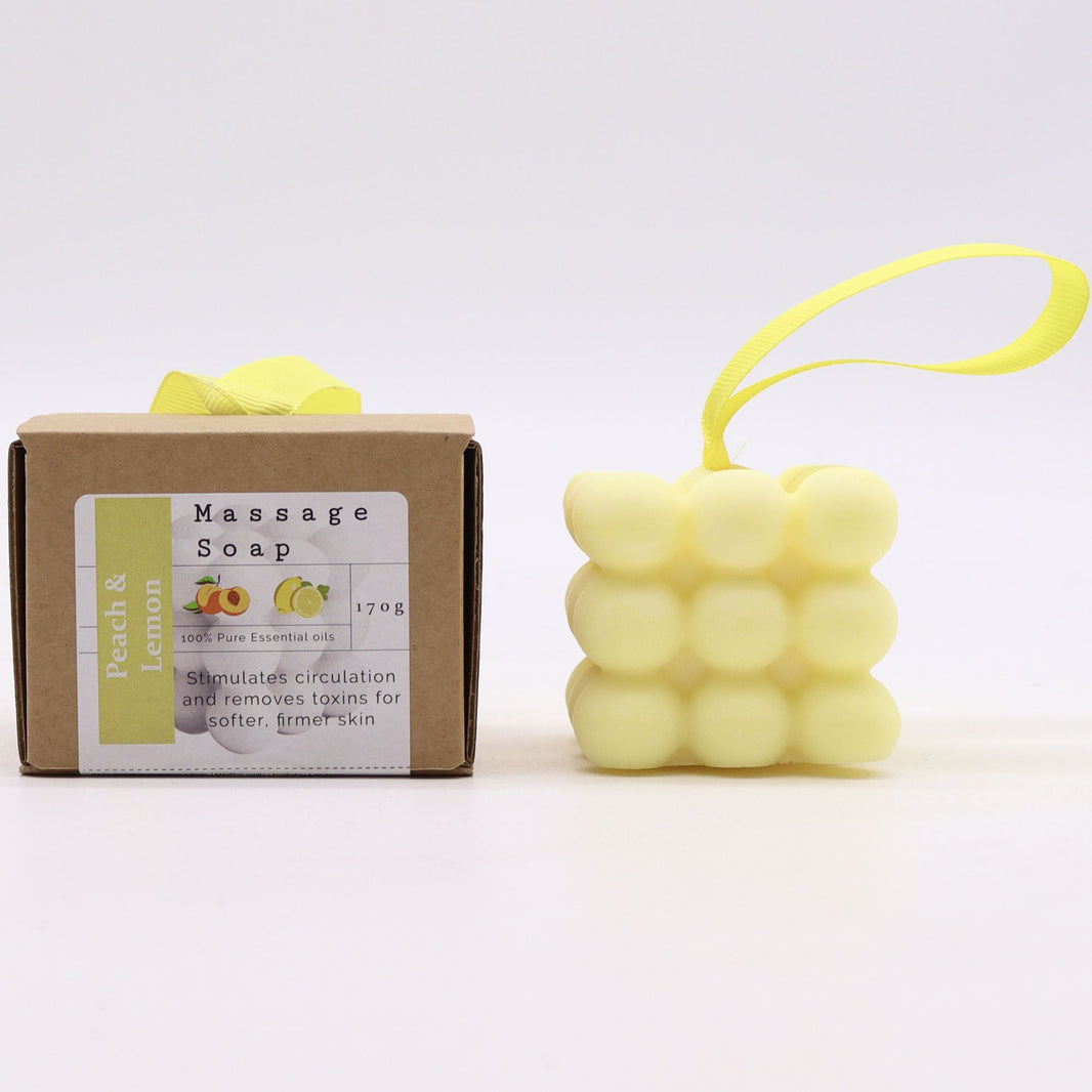 A pale yellow square massage soap bar with raised nodules for skin stimulation, housed in a white gift box with an orange label.