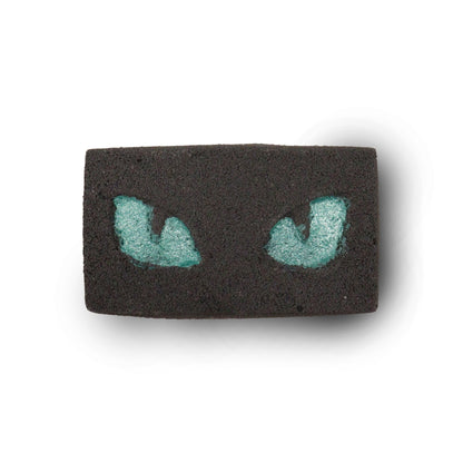 A dark, charcoal-grey bath bomb featuring two glowing-style green cat eyes, creating a mysterious and fun bath experience.