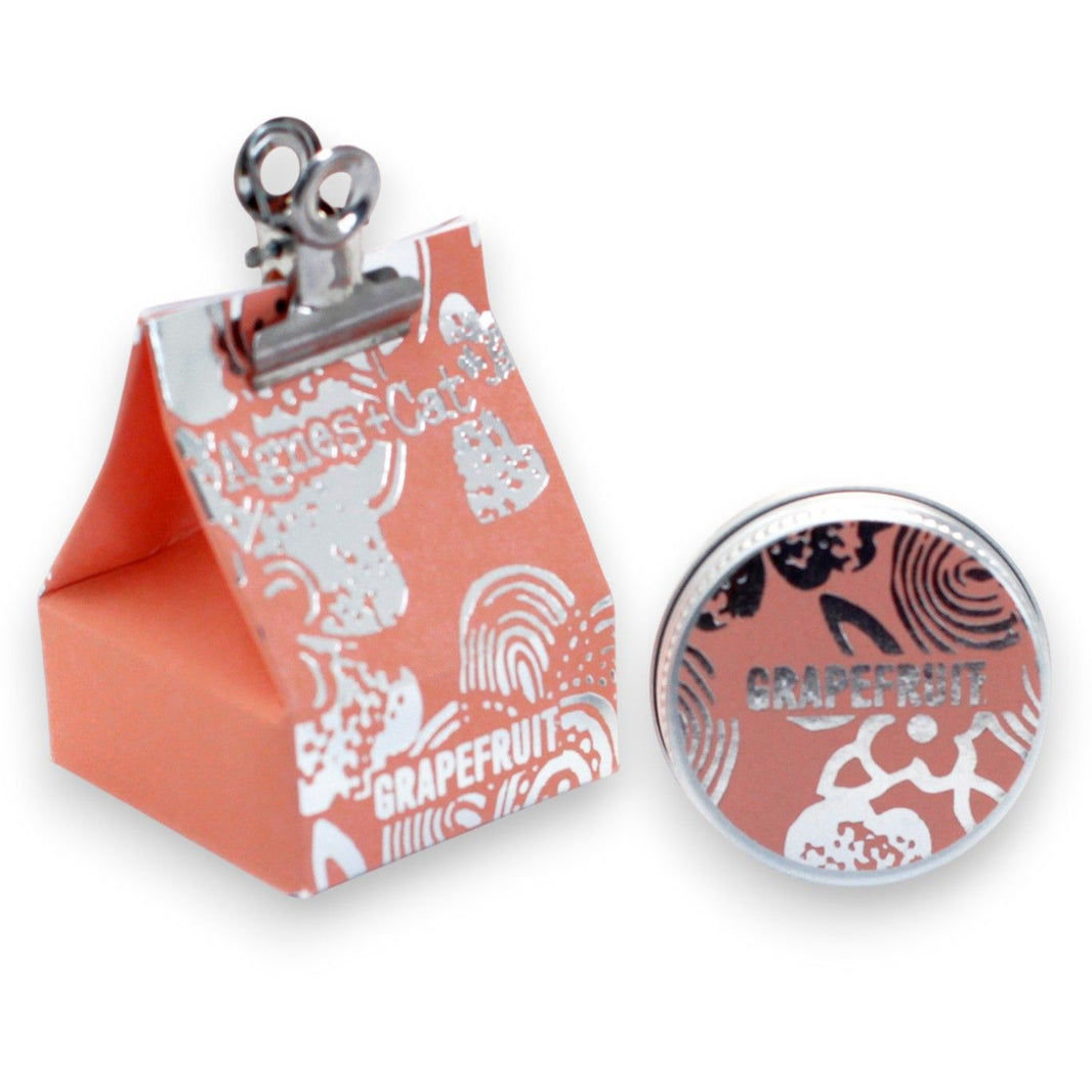 A 20g round tin of grapefruit-scented lip balm shown with its bright orange-themed gift packaging.