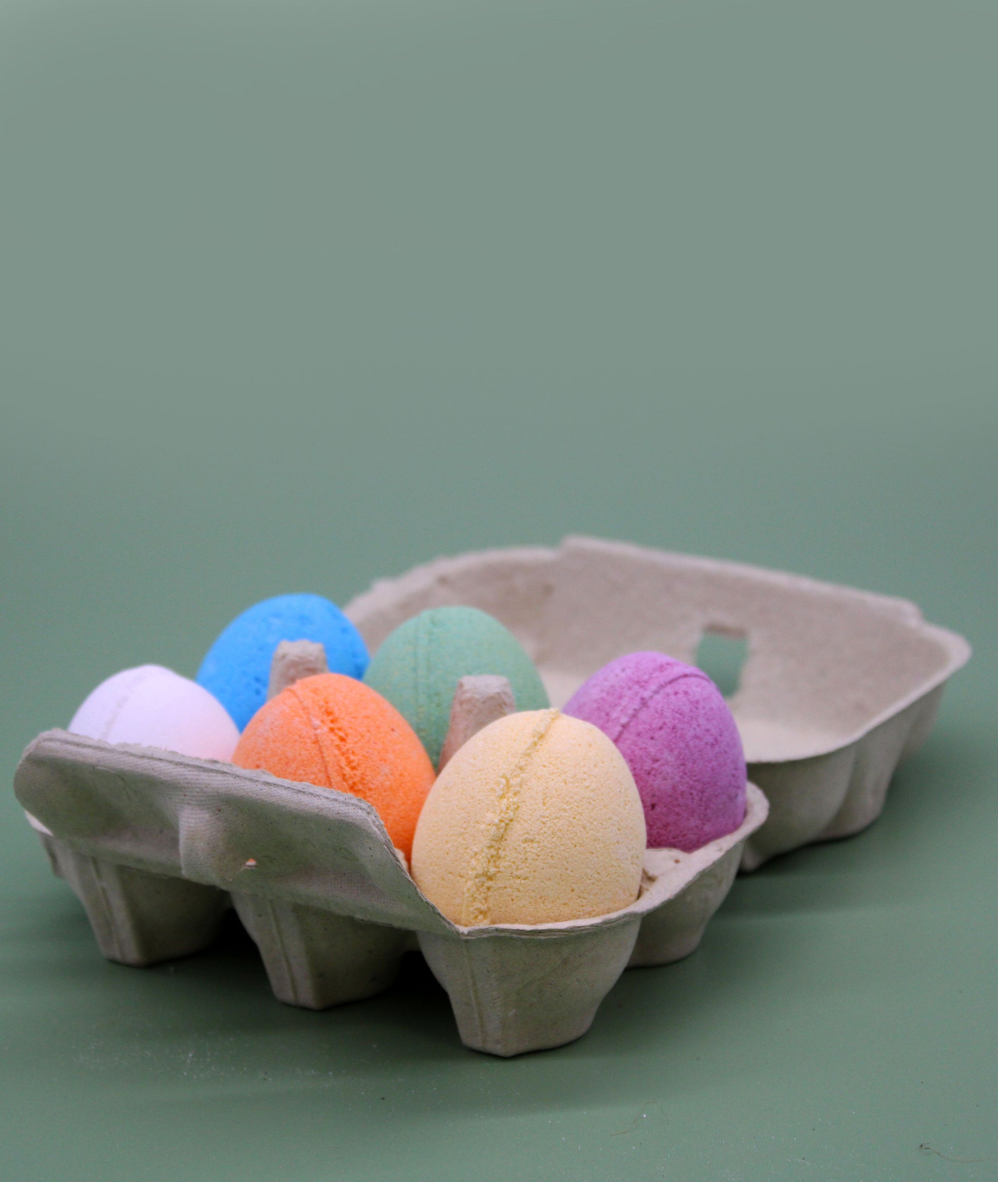 A cardboard egg carton filled with six different colored egg-shaped bath bombs, perfect for a Spring or Easter gift set.