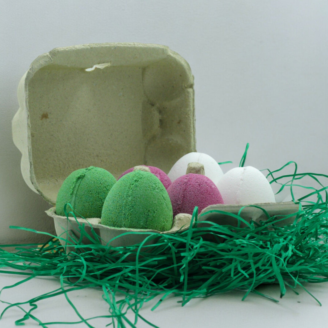 A cardboard egg carton holding 6 colorful, egg-shaped bath bombs in various Spring shades like green, pink, and white.