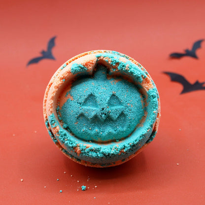 A bright orange, pumpkin-shaped bath bomb with a carved jack-o'-lantern face detail and a green stem.